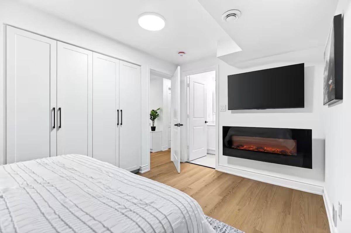 Modern bedroom with white built-in wardrobes, wooden flooring, bed, wall-mounted TV, and electric fireplace insert.