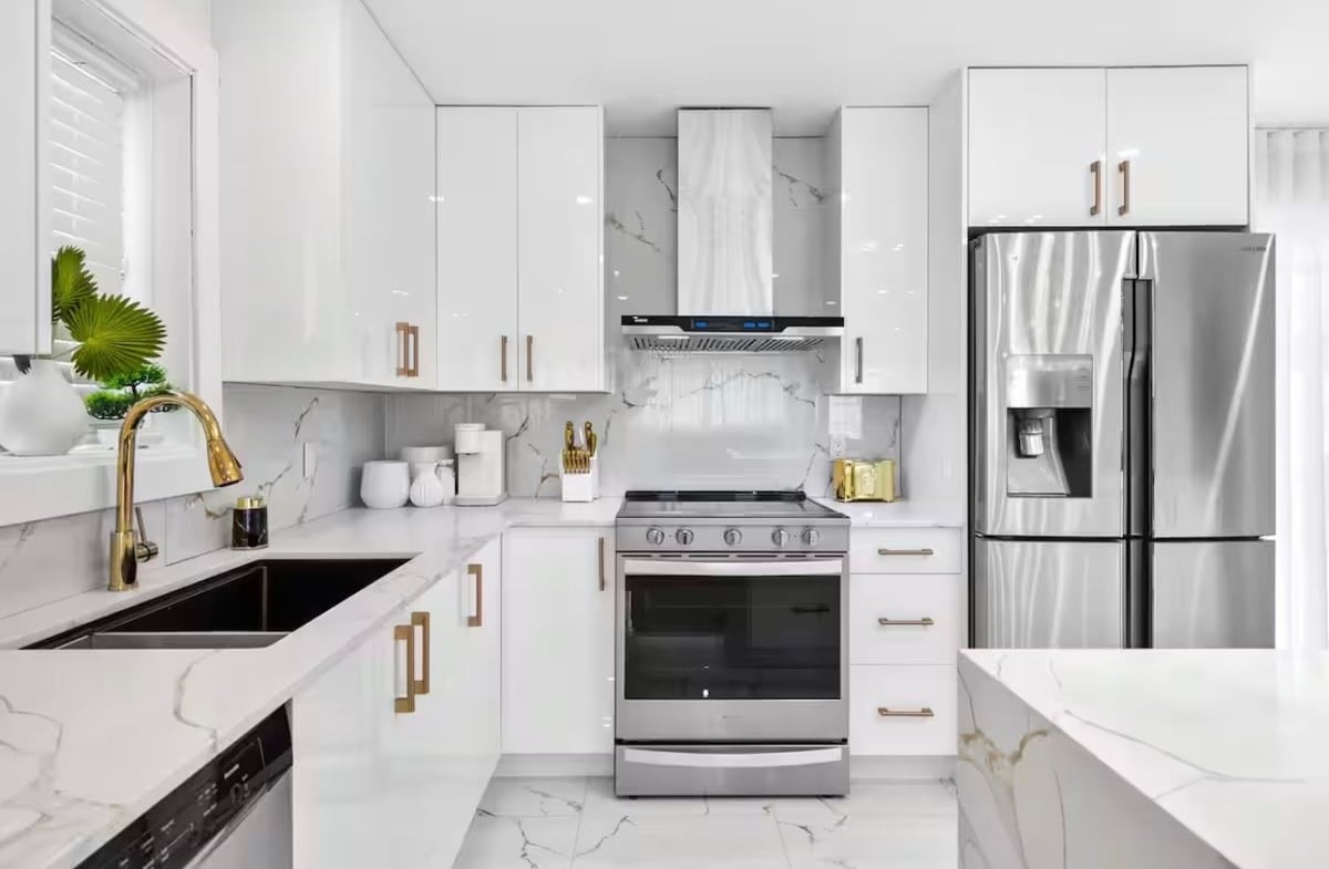 Modern white kitchen with stainless steel appliances, marble countertops, and gold hardware accents